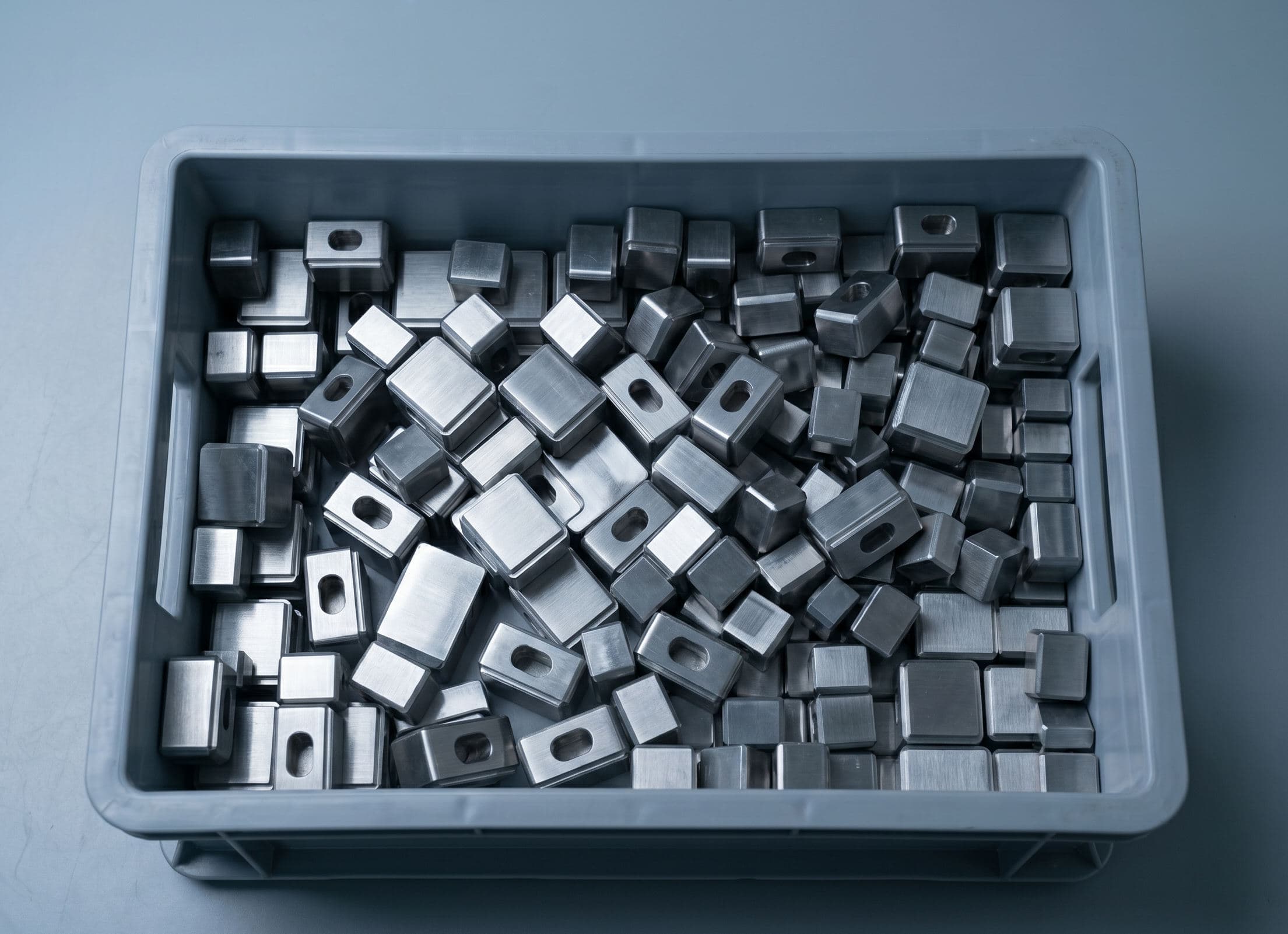 Gray industrial parts tote holding unmarked CNC-machined steel parts, staged on a work table before laser marking.
