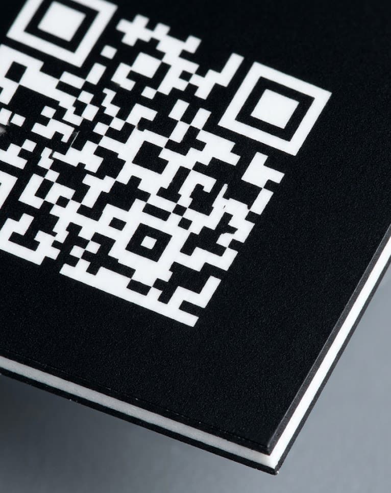 Cross-section detail of Tesa 6973 — black matte top layer with the white sub-layer revealed where the laser has bleached a high-contrast Data Matrix code. Tight macro lighting, no clutter.