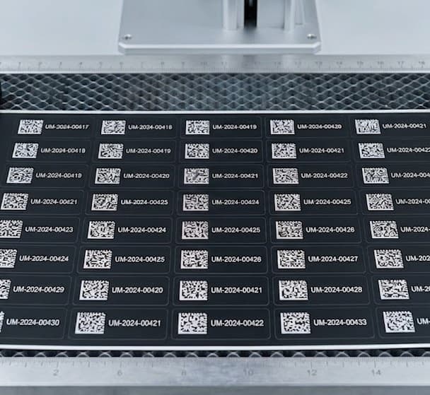Sheet of Tesa 6973 black-matte polyacrylic on the laser bed mid-process — kiss-cut grid of asset tags, each marked with a unique QR code and serial in crisp white-on-black.