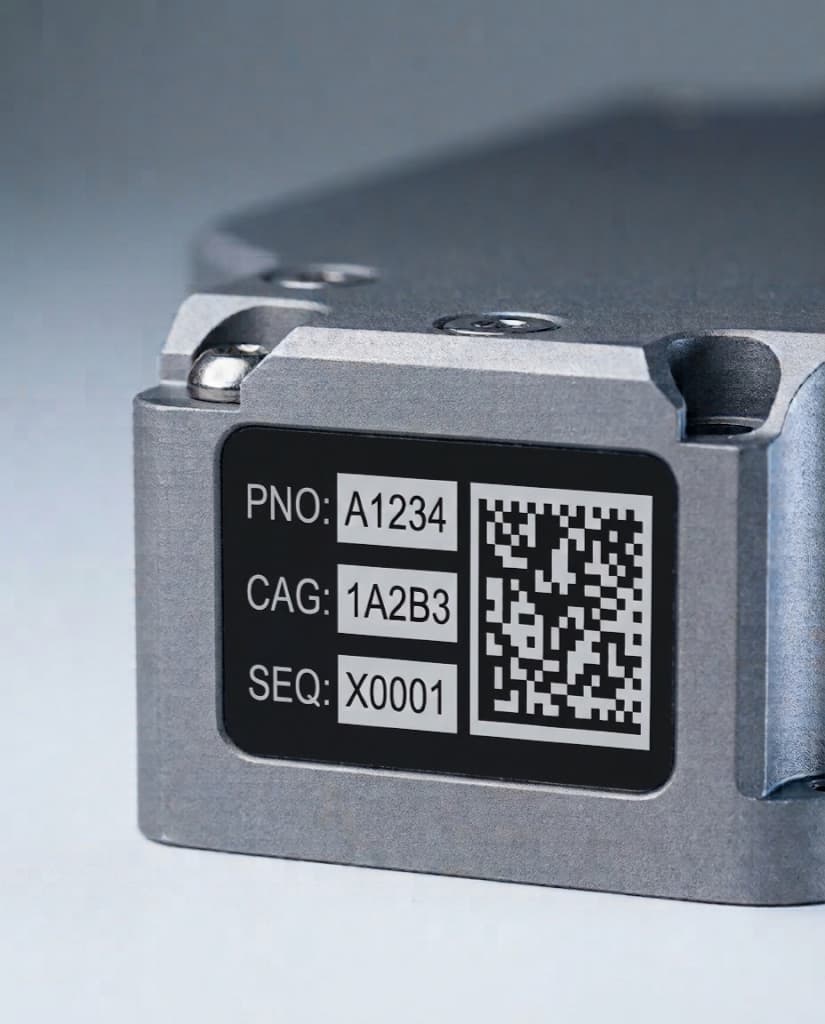 Macro of a finished MIL-STD-130 UID label on black-matte acrylate stock, applied to the end face of a machined aluminum housing: human-readable PNO, CAG, and SEQ data fields at left, ECC 200 Data Matrix at right.